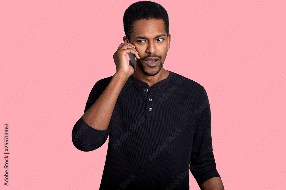 African American man has telephone coversation, hears something unpleasant news, looks aside, wears black outfit. Handsome dark skinned guy holds cell phone in hand isolated over rose background. African American man has telephone coversation, hears something unpleasant news, looks aside, wears black outfit. Handsome dark skinned guy holds cell phone in hand isolated over rose background.