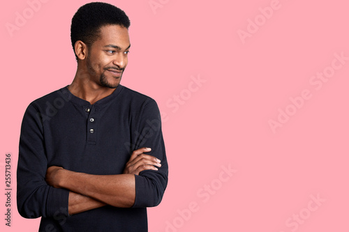 Portrait of confident charismatic young handsome African American male in black casual shirt with hands crossed on chest. Guy looks smiling aside. Attractive man with short hairstyle and beard poses.