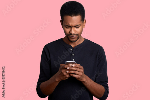 Young african man with mobile phone over rose background. Handsome dark skinned male types massege with bad news for his boss. Attractive black guy dresses casual outfit poses in photo sstudio. © sementsova321