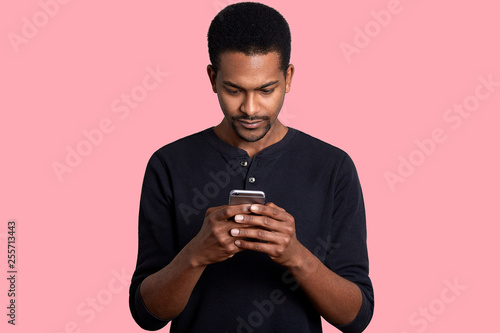 Young guy looks at the screen of smartphone with serious fecial expression. Black handsome male has important unpleasent correspondence. Boring pastime. Modern technology and Internet concept. photo