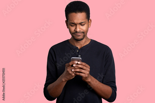 Attractive black man holds mobile phone and looks worried. Black guy reads message with unpleasent news from friend and does not know how to help. Male wears casual jumper, stands against pink wall. © sementsova321