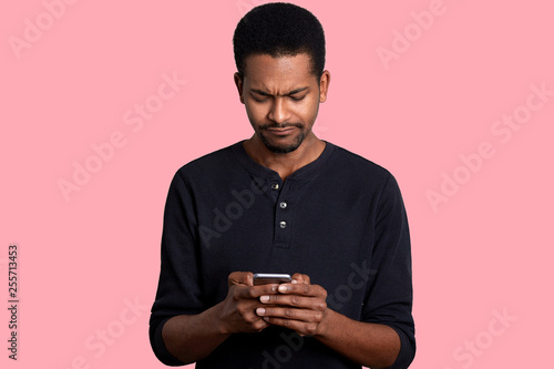 Portrait of attractive African American man with telephone in hands, surfes Internet. Handsome black male looks weather forecast for weekend with sad fecial expression. Modern technology concept. © sementsova321