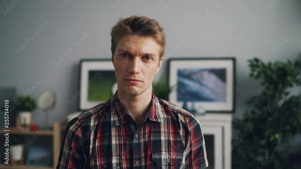 Portrait of handsome young man looking at camera with unhappy face wearing bright checkered shirt standing in modern apartment alone. Youth and emotions concept. Portrait of handsome young man looking at camera with unhappy face wearing bright checkered shirt standing in modern apartment alone. Youth and emotions concept.