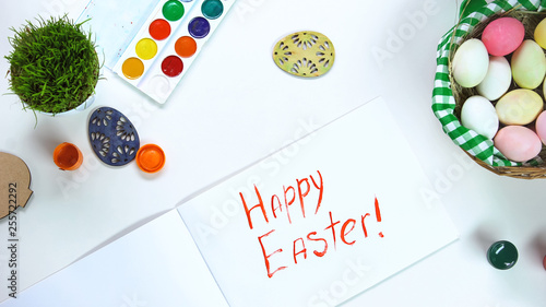 Happy Easter phrase written on paper, table with decorative eggs and paints