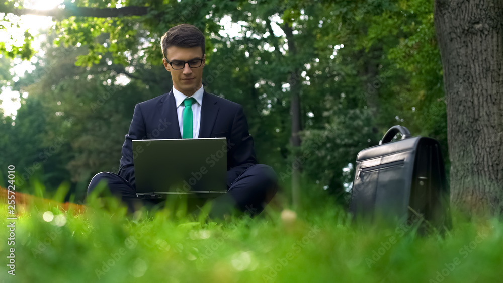 Business man sitting on grass and working on laptop in park outside office Business man sitting on grass and working on laptop in park outside office