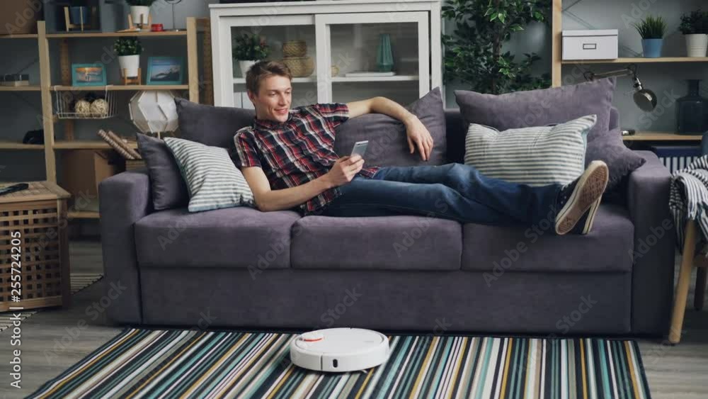 Happy student is using smartphone looking at screen and smiling relaxing on comfortable couch while robotic vacuum cleaner is vacuuming carpet and floor.