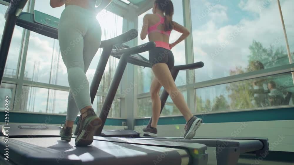 Two fit girls running on treadmill exercise machine in sunlight to warm-up before gym workout