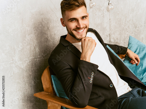 Portrait of handsome smiling hipster lumbersexual businessman model wearing casual black suit. Fashion stylish man posing sitting in chair in luxury interior. Thinking photo