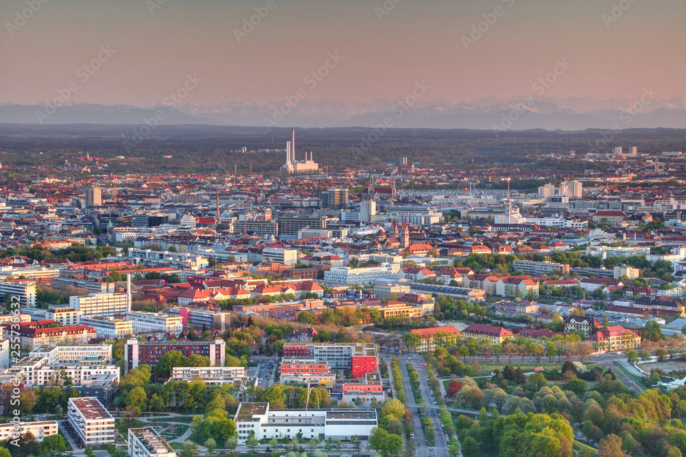 Aerial view of modern European city outskirts in evening sunlight with tower blocks, chimneys, industrial, commercial and office buildings and Bavarian Alps in background, Munchen Bayern Germany
