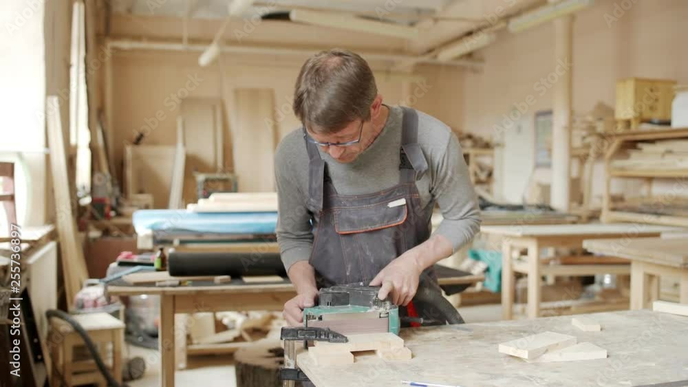 Lockdown medium shot of mature joiner in overalls using belt sander to sand a piece of wood attached to workbench in his workshop Lockdown medium shot of mature joiner in overalls using belt sander to sand a piece of wood attached to workbench in his workshop