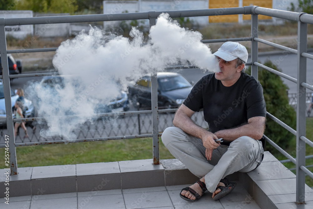 Elderly man hold and smoke his electronic cigarette outdoor among parking lot and cityscape. man really likes process of smoking Elderly man hold and smoke his electronic cigarette outdoor among parking lot and cityscape. man really likes process of smoking