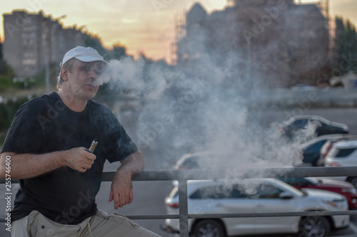 Elderly man hold and smoke his electronic cigarette outdoor among parking lot and cityscape. man really likes process of smoking