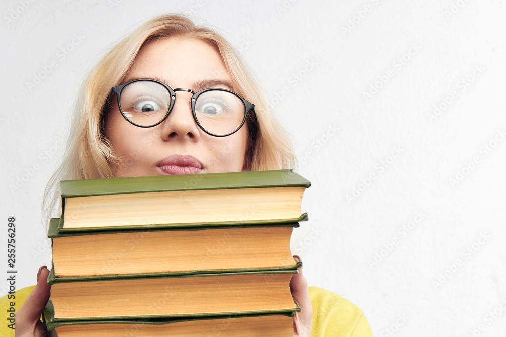 Funny clever girl looks out of a pile of books. Blonde nerd in round glasses on a white background. Educational concept Funny clever girl looks out of a pile of books. Blonde nerd in round glasses on a white background. Educational concept
