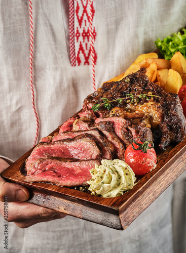 Male hands holding a wooden board with a juicy steak with fragrant butter. Sliced Ribeye Steak with Potatoes, Onions and Baked Cherry Tomatoes