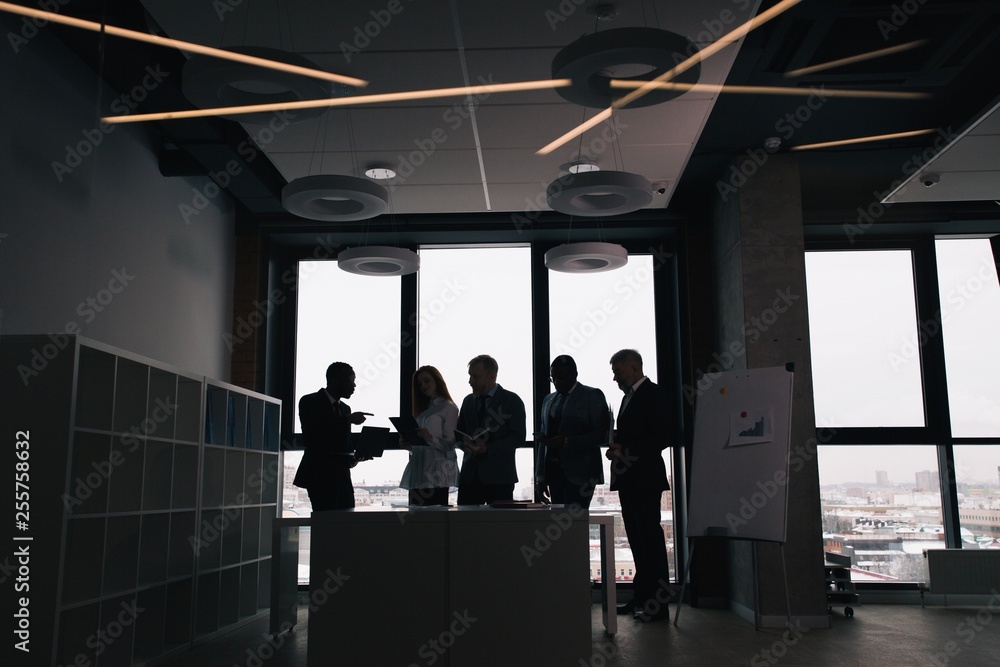 Corporate business team working in the office space standing at the table. Dark photo silhouettes of businessmen on the background of the window Corporate business team working in the office space standing at the table. Dark photo silhouettes of businessmen on the background of the window