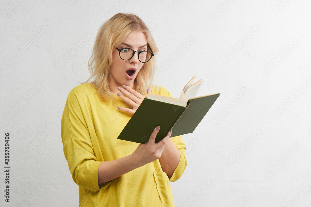 Young blonde in round glasses reads a book with a surprised face in yellow clothes. Isolated in white studio. Young blonde in round glasses reads a book with a surprised face in yellow clothes. Isolated in white studio.