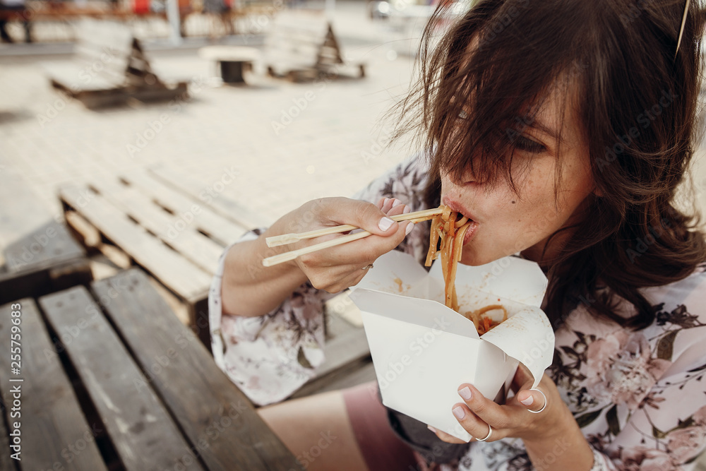Stylish hipster girl eating wok noodles from box with bamboo chopsticks. Asian Street food festival. Boho woman eating thai noodles takeaway paper box. Food delivery Photos | Adobe