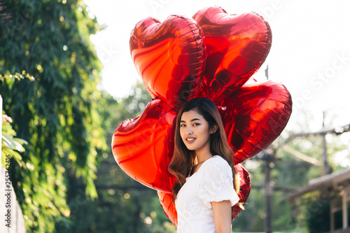 Pretty Asian woman with bunch of red heart shaped balloons looking away while standing on blurred background of park on sunny day