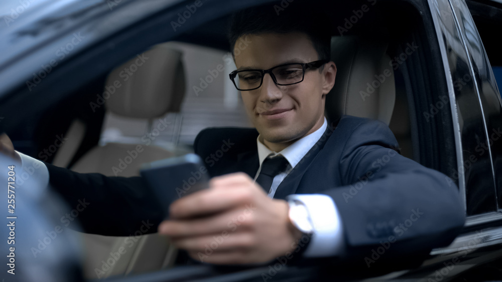 Glad businessman texting with girlfriend and smiling, sitting in car, smartphone