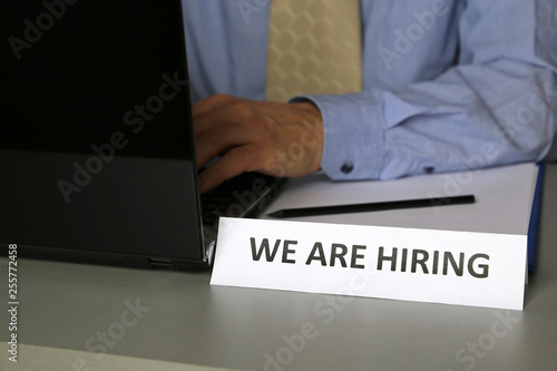 Hiring and recruitment, man in blue shirt and tie sitting at desk with laptop near the sign with the inscription We are hiring. Concept of headhunter, office manager, staffing, personnel officer