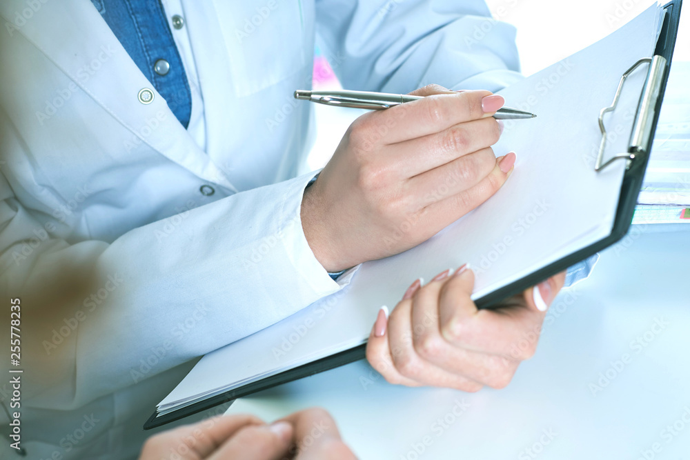 Female doctor arm hold silver pen filling patient complaints list clipped to pad. Female patient tells the doctor about her problems. Physical exam or disease prevention concept. Female doctor arm hold silver pen filling patient complaints list clipped to pad. Female patient tells the doctor about her problems. Physical exam or disease prevention concept.