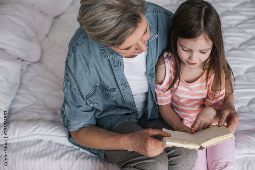 Gray-haired woman teaching her granddaughter to read book at home Gray-haired woman teaching her granddaughter to read book at home