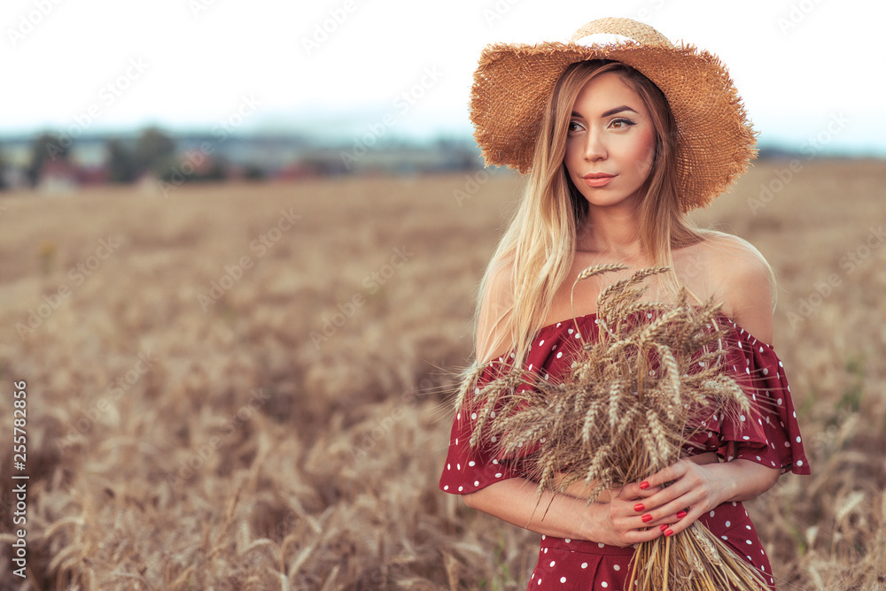 Obraz premium Beautiful girl red dress straw hat. In summer wheat field. Free space for text. Harvesting in village, wheat ears in hand. Long hair tanned skin, emotions of tenderness confident look. Place for text.