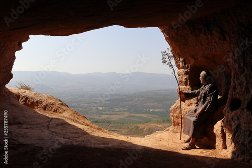 Billede på lærred HERMIT WITH A TREE IN THE HAND SEATED IN A CAVE ON THE HIGH OF A VALLEY