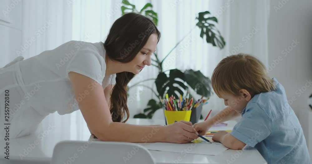 Young beautiful mother and son draw with colored pencils sitting at the table in the kitchen. The camera moves in slow motion Young beautiful mother and son draw with colored pencils sitting at the table in the kitchen. The camera moves in slow motion