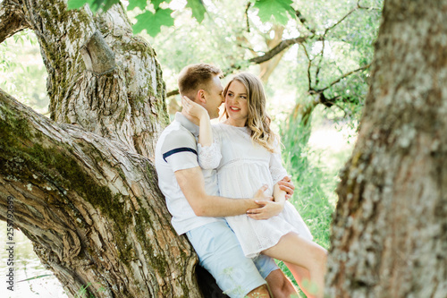 Young pretty couple in love lying on tree in park. Handsome cheerful blonde girl in white dress hugging her boyfriend. Man and woman having fun outdoors