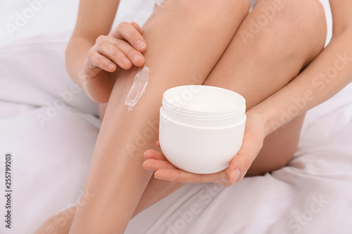 Young woman applying cream on her legs on bed, closeup. Beauty and body care