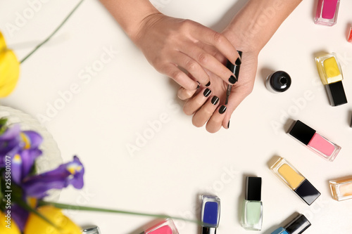 Woman applying nail polish near bottles on white table with flowers, top view
