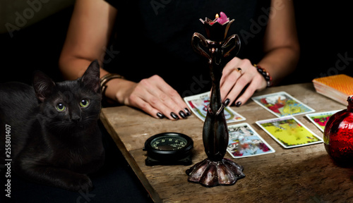 The fortune teller lays out on a wooden table the tarot cards by the light of a candle. Black cat sitting near the table. Selective focus. photo