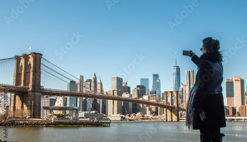 Photography Young Woman tourist by the river in Dumbo taking pictures of Brooklyn Bridge and Cityscape of New York skyline
