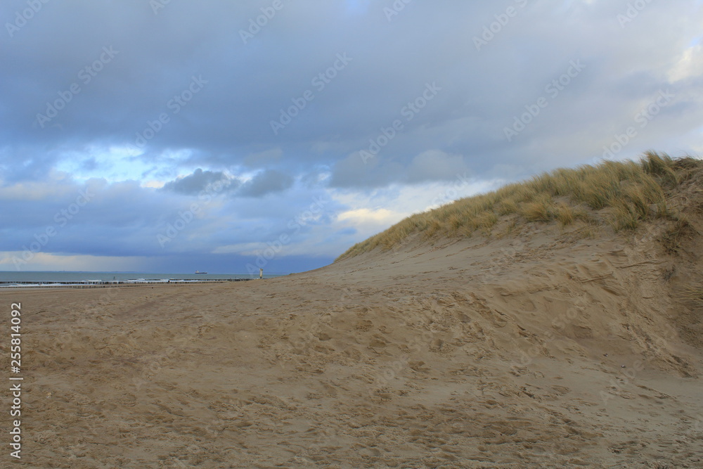 big sand dune with grass and the sea in the background in zeeland, holland in winter with a stormy blue sky with clouds