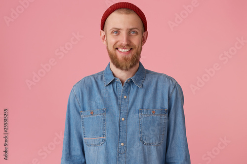 Glad joyful guy in red hat with red thick beard smiling happily showing white healthy teeth, wears fashionable denim shirt, isolated on pink background