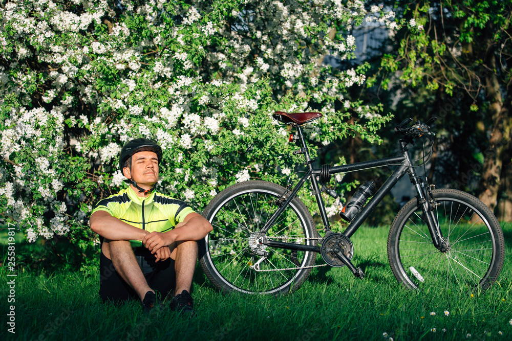 Professional cyclist in helmet and spotswear sitting with mountain bicycle near blooming tree. Young rider resting in park after ride.