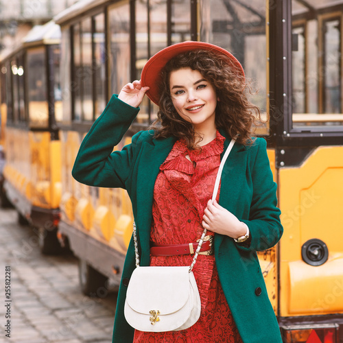 Outdoor portrait of young beautiful happy smiling lady wearing orange hat, snakeskin printed dress, green coat, with white cross body bag, posing in street of European city photo