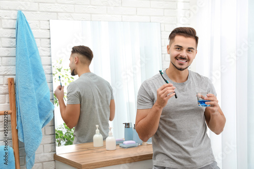Young man with toothbrush and glass of mouthwash in bathroom at home, space for text. Teeth and oral care