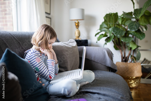 female child sitting on the sofa at home with a laptop and head phones