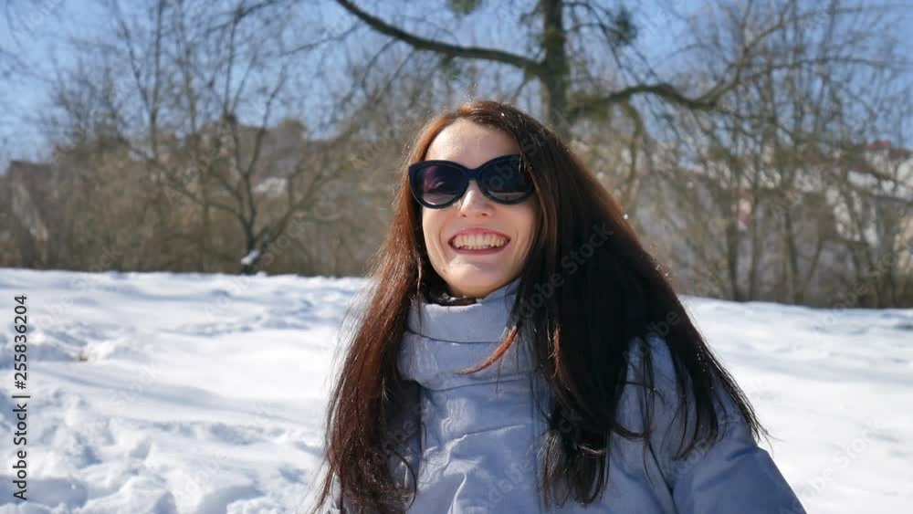 Active smiling woman in dark sunglasses is playing snowballs on blue sky background in the park during sunny day. Outdoors activities concept Active smiling woman in dark sunglasses is playing snowballs on blue sky background in the park during sunny day. Outdoors activities concept