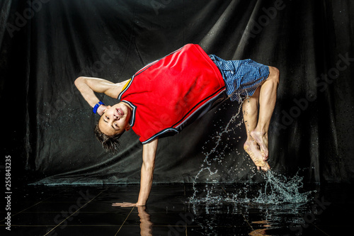 cool and happy korean young guy break dancer in style of bboying doing complex tricks on floor with water splashes and liquid drops effects in aqua zone photo studio with black isolated background photo