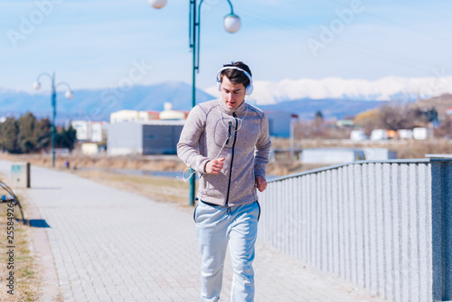 Fototapeta Naklejka Na Ścianę i Meble -  A panting athlete running fast on a track while wearing headphones and grey sweatpants, early in the morning on a sunny day..