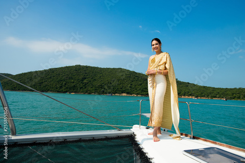 Asian Woman in Gold Yellow Thai Traditional Costume with jewelry accessories get Winds on Luxury Yacht boat ship in Green Ocean nice sky, Lady relaxing on cruise wearing old formal dress