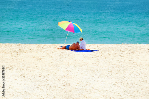 Fototapeta Naklejka Na Ścianę i Meble -  Happy Senior Couple on a tropical beach relax in the sun on sand under a red umbrella. Travel background .