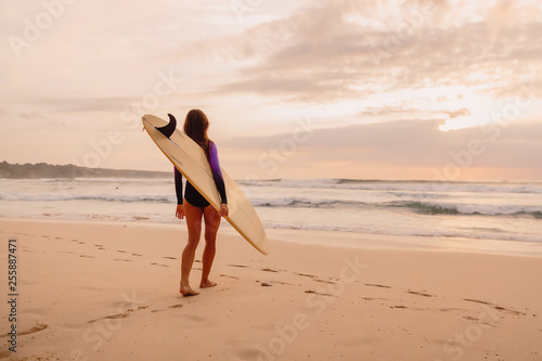 Surfer girl with surfboard on a beach at sunset or sunrise.