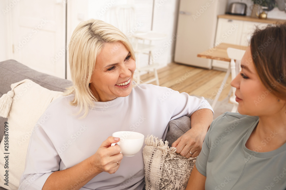 Best friends with my mom. Pretty girl smiling while talking to her mother sitting on cozy couch, drinking coffee and sharing secrets. Family, parenting, togetherness and relationships concept Best friends with my mom. Pretty girl smiling while talking to her mother sitting on cozy couch, drinking coffee and sharing secrets. Family, parenting, togetherness and relationships concept