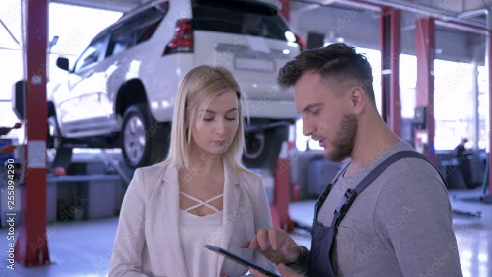 car repair shop, mechanic with digital tablet talking to a female client on background of the automobile on hydraulic lift car repair shop, mechanic with digital tablet talking to a female client on background of the automobile on hydraulic lift