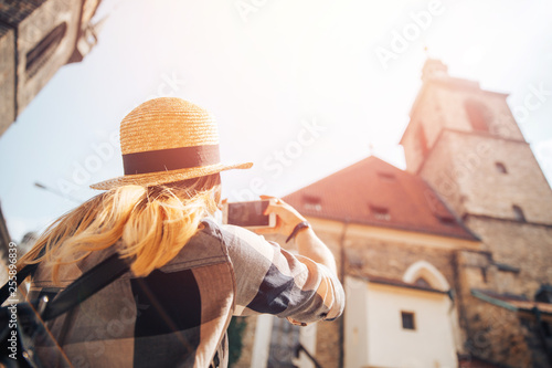 Blonde girl in straw hat, plaid shirt takes photo on phone of European stone buildings medieval architecture © Parilov