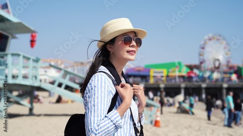 young asian girl backpacker with straw hat standing on beach bay on Santa Monica Pier near safeguard station. female photographer sightseeing ocean view. colorful amusement park with ferris wheel.